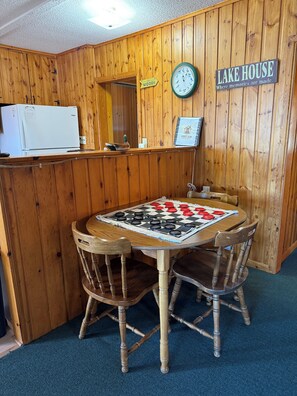 A wooden interior with a checkered table, wooden chairs, a clock, and a sign reading 'LAKE HOUSE.'