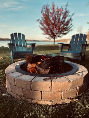 A circular brick fire pit with logs burning, surrounded by grass, with two Adirondack chairs and a tree in the background. - The Cozy Cub (Crosslake)