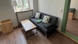 A room with a wooden floor, a sofa with cushions, a table, a window with a view, and a radiator. - Fichtelzimmer - central and modern vacation apartment in the heart of Wunsiedel (Wunsiedel)