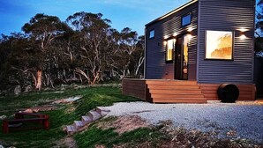Una cabaña moderna de dos pisos con terraza de madera, ubicada en un entorno natural con árboles al fondo.