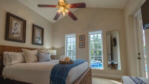 Ein geräumiges Schlafzimmer mit einem großen Bett, einem Deckenventilator, gerahmten Kunstwerken und Blick auf einen Swimmingpool.