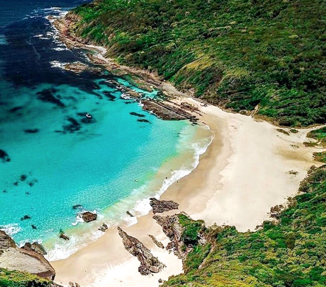 An aerial view of a coastal area with a sandy beach, clear turquoise waters, and lush green vegetation.