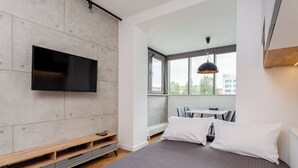 Une chambre moderne avec un grand lit, un téléviseur fixé au mur, une fenêtre donnant sur la ville et une petite table avec des chaises.