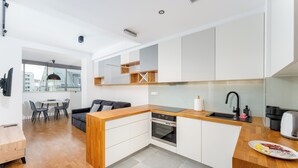 Une cuisine moderne avec des armoires blanches, un plan de travail en bois et un four encastré. On y trouve un coin repas avec une table et des chaises, ainsi qu'un canapé à l'arrière-plan.
