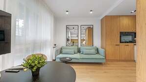 A modern living room with a sofa, a dining table with a plant, and a kitchen area with wooden cabinets and an oven. - Kluczborska Chic Studio (Kraków)