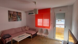 Una sala de estar con un sofá, una mesa de centro y dos sillones. Se ve una ventana con persianas rojas y una puerta que da al exterior.