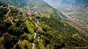 Vista aérea de un pueblo montañoso con un camino sinuoso, abundante vegetación y un pequeño cuerpo de agua.