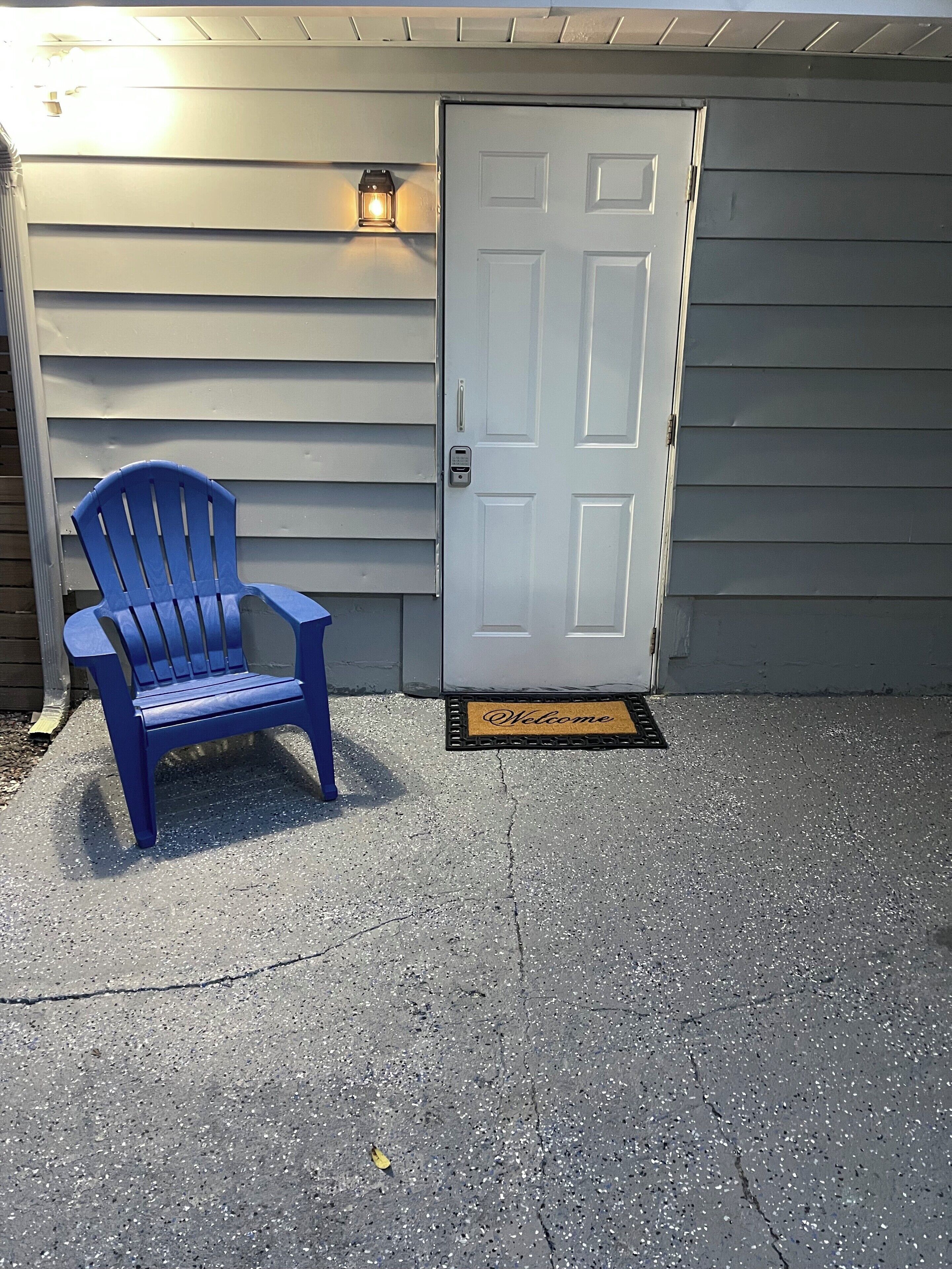 Una silla Adirondack azul frente a un edificio con una puerta blanca y un felpudo.
