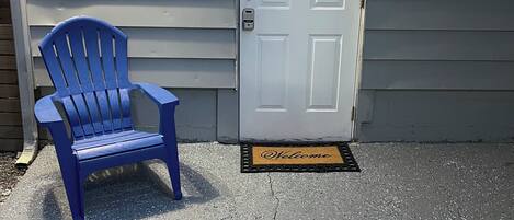 Una silla Adirondack azul frente a un edificio con una puerta blanca y un felpudo.