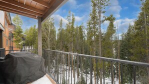 A balcony with a metal railing, a covered grill, and a view of a forest with tall pine trees.