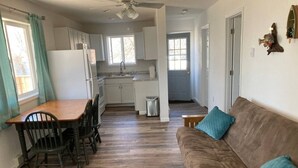 A spacious living area with a wooden dining table and chairs, a brown sofa with teal cushions, and a kitchen in the background.