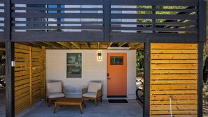 Un espace extérieur aménagé, abrité et doté d’un canapé et d’une table, une porte avec un cadre orange et une clôture en bois.