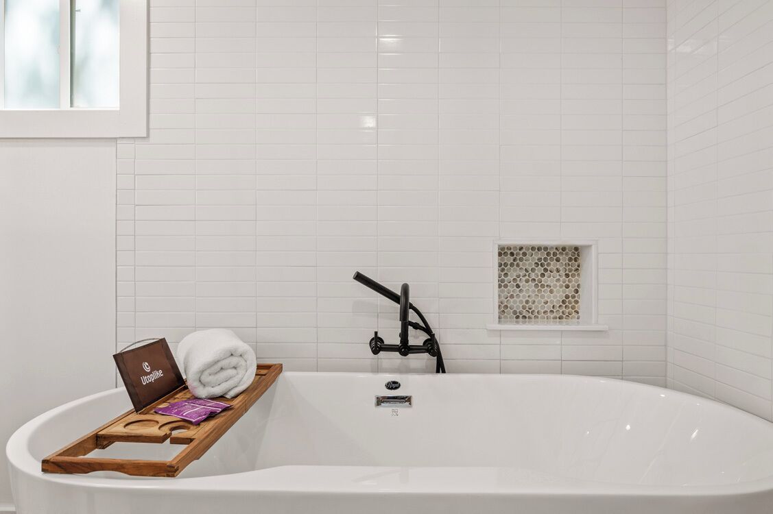A modern bathroom with a white bathtub, a wooden tray with toiletries, and a small window with a honeycomb pattern.