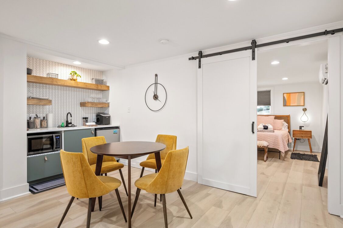 A modern kitchen with a microwave, oven, and wooden shelves, a dining area with a round table and yellow chairs, and a bedroom with a bed and bedside table.