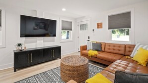 Un salon moderne avec un meuble de rangement noir pour les appareils électroniques, un canapé en cuir marron et un pouf en fibres tressées.