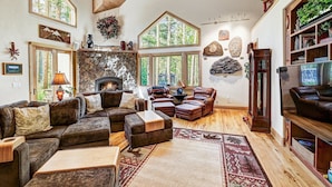 A cozy living room with a stone fireplace, leather furniture, and large windows offering views of the outdoors. - NEW Cedar Lodge on Peak 8 | Private Hot Tub, Firepit, Grill & Easy Shuttle Access (Breckenridge)