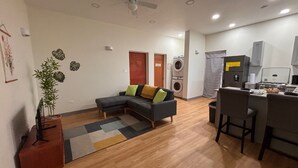 Sala de estar con sofá modular, una mesita y una planta. Se observa una cocina con refrigerador, estufa y pileta. Se puede ver un ventilador de techo.