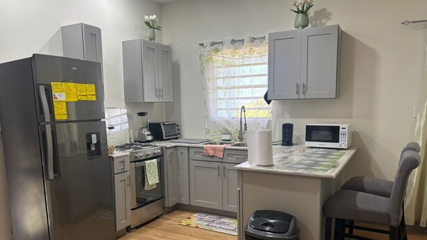 Una cocina compacta con gabinetes grises, un refrigerador de acero inoxidable, un microondas, una estufa y un cesto de basura.