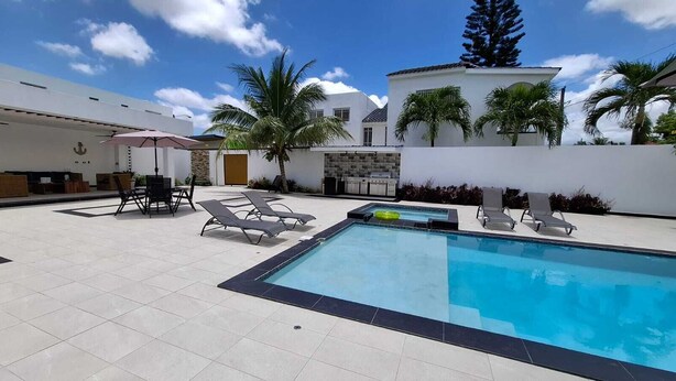 A swimming pool with lounge chairs and a table, surrounded by a white wall and palm trees.