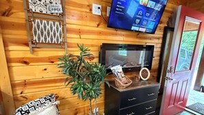 Una habitación acogedora con paredes de madera, un televisor de pantalla plana sobre la chimenea, una planta pequeña y un escritorio con cajones.