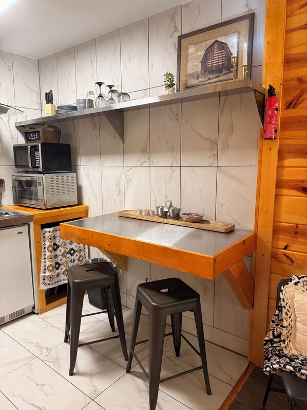 A compact kitchen with a wooden countertop, two stools, a microwave, a toaster, and a small shelf with a framed picture.