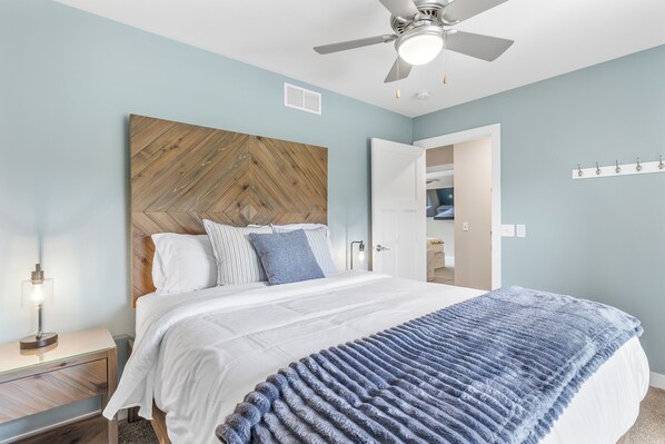 A bedroom with a wooden headboard, a bed with white bedding, a blue blanket, a nightstand with a lamp, and a ceiling fan.