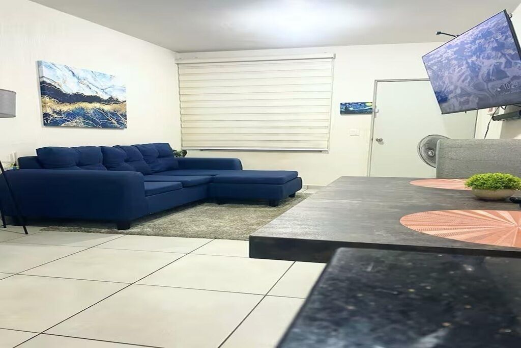 A living room with a blue sectional sofa, a black coffee table, and a flat-screen TV mounted on the wall.