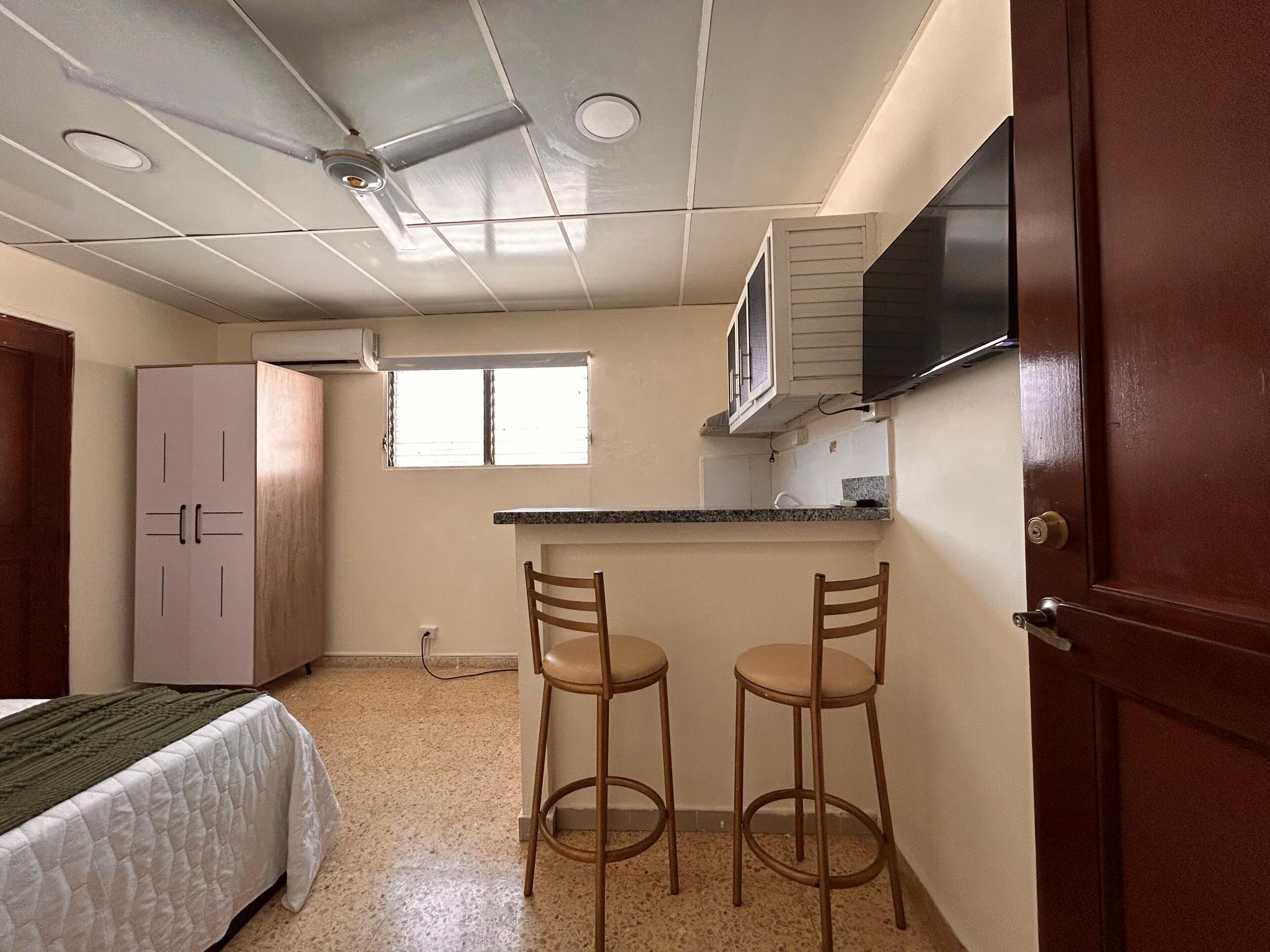 Habitació petita amb un llit, una cuineta, dos tamborets i un televisor fixat a la paret.
