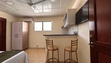 Habitació petita amb un llit, una cuineta, dos tamborets i un televisor fixat a la paret.