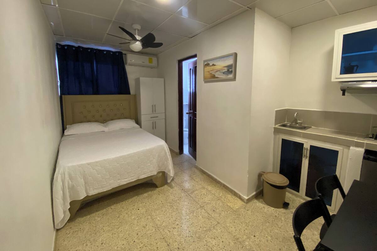 A bedroom with a bed, a ceiling fan, a window with curtains, a door, a small kitchenette, and a trash bin.