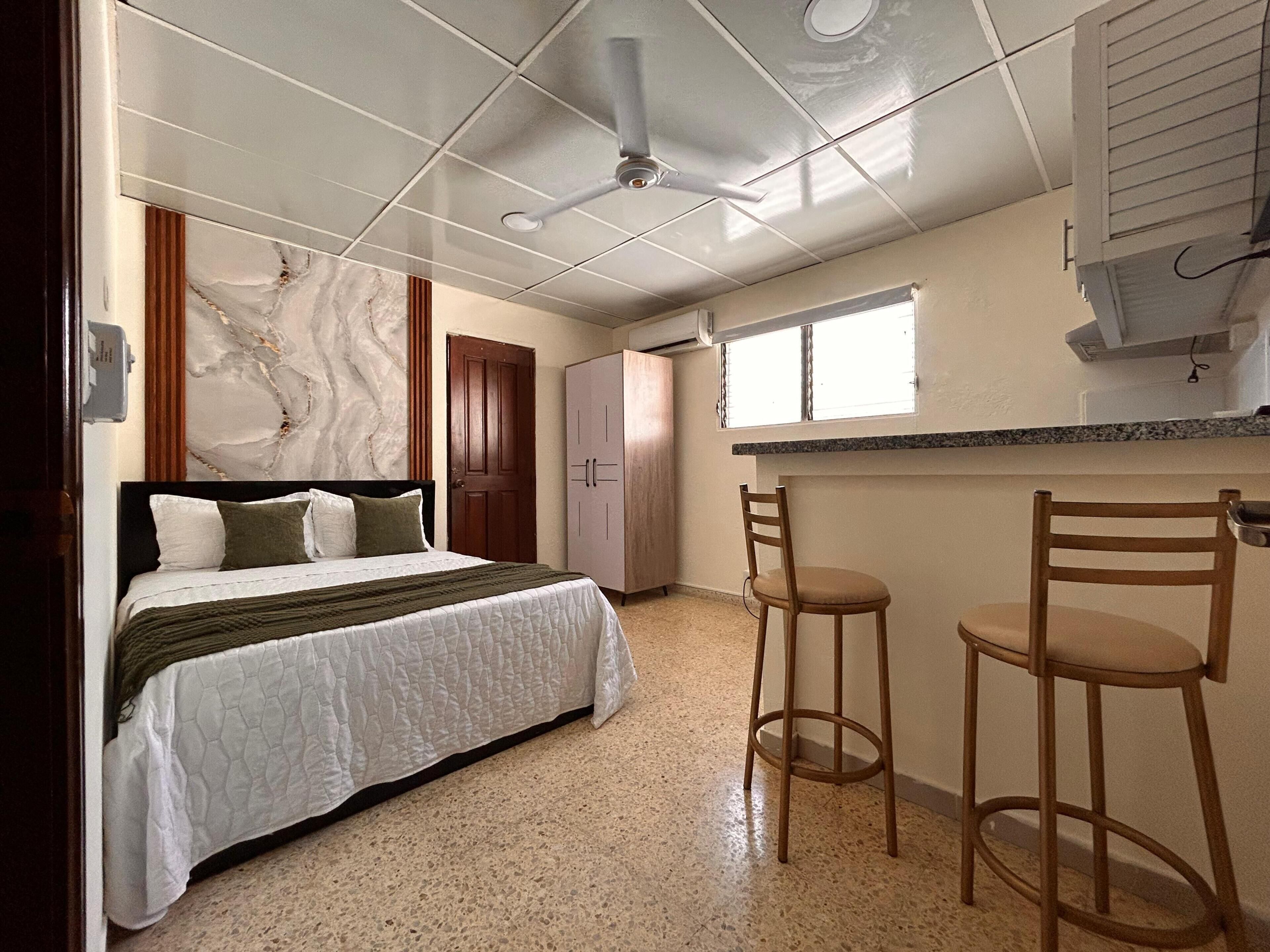Quarto com cama, ventoinha no teto, parede de mármore, porta de madeira e área de cozinha compacta com janela.