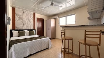 Quarto com cama, ventoinha no teto, parede de mármore, porta de madeira e área de cozinha compacta com janela.