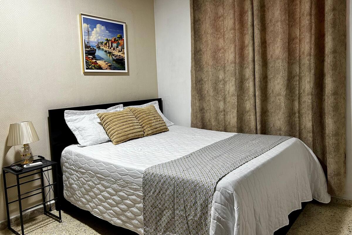 A neatly made bed with a quilted cover, a framed picture on the wall, and a curtain on the window.