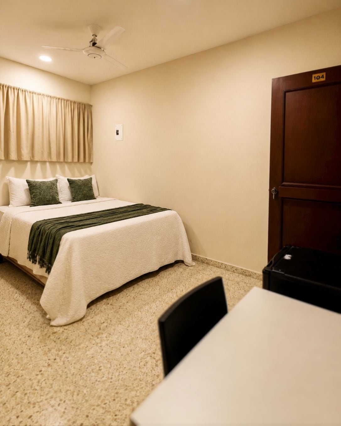A hotel room with a bed, a desk, a chair, a ceiling fan, and a door.