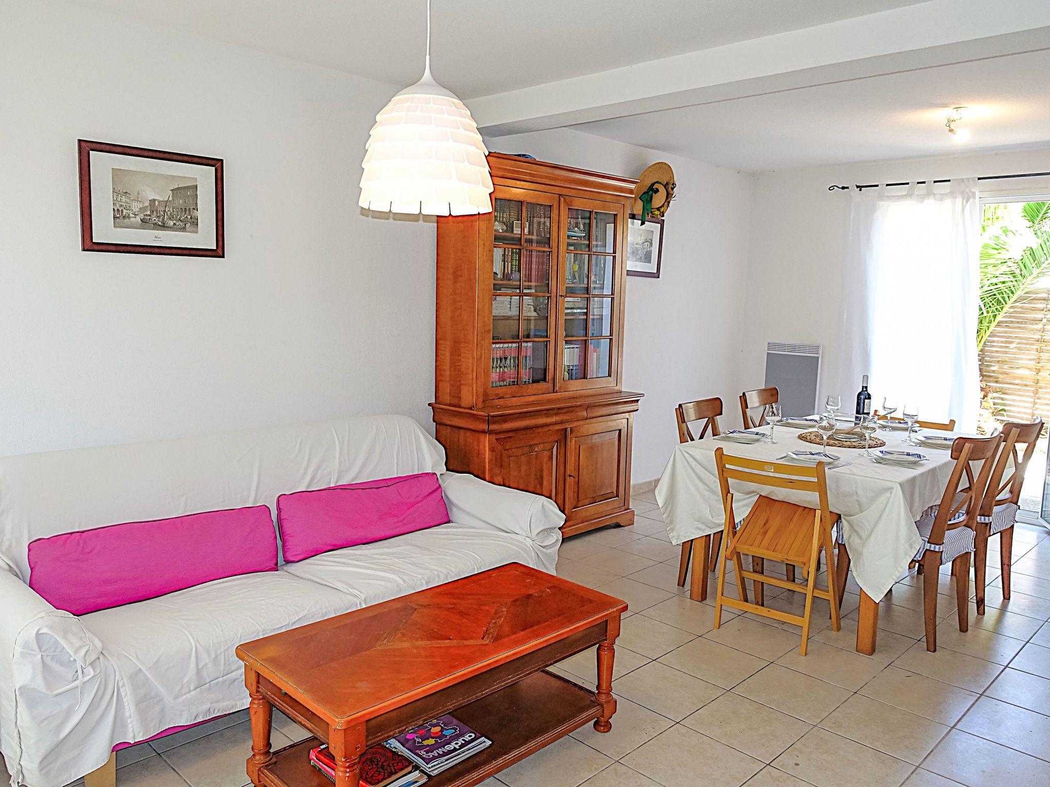 A living room with a white sofa, a wooden coffee table, a dining area with a table set for four, and a wooden cabinet with glass doors.