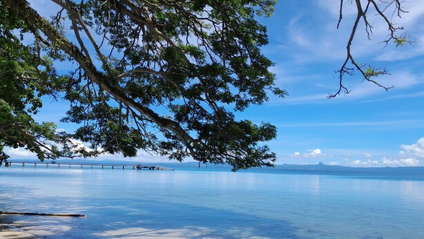 A beach with clear water, a wooden pier, and a distant mountain range.