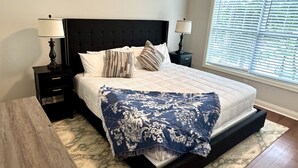 A bedroom with a large bed, two bedside tables, a nightstand, a window with blinds, and a patterned rug. - American Pharoah Cottage | 3BR Downtown Versailles (Versailles)