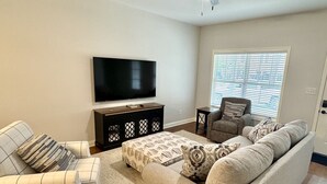 A living room with a sofa, ottoman, and armchair, a flat-screen TV, and a window with blinds. - American Pharoah Cottage | 3BR Downtown Versailles (Versailles)