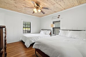 A bedroom with two beds, wooden flooring, white walls, a ceiling fan, and windows with blinds.