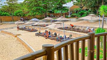 A beachside deck with lounge chairs and umbrellas, surrounded by lush greenery and a sandy beach area.