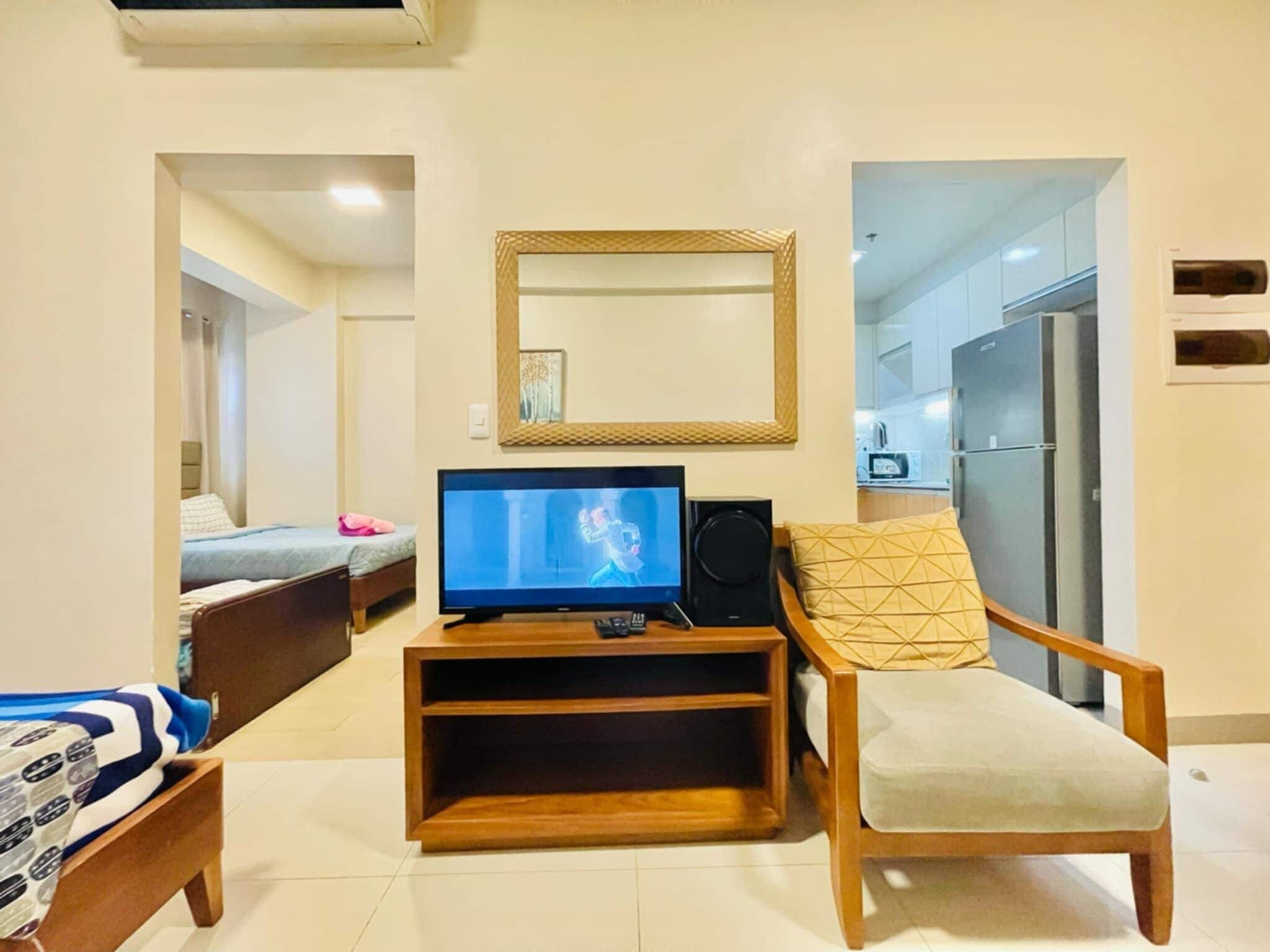 A hotel room with a bed, a TV, a wooden side table, a chair, a mirror, and a kitchenette.