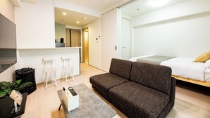 A modern, well-lit room with a sofa, a small table, and a bed. There are white chairs and a counter in the background. - Hotel Reference Ropponmatsu Condominium (Fukuoka)