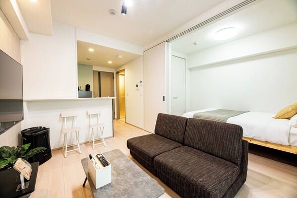 A modern, well-lit room with a sofa, a small table, and a bed. There are white chairs and a counter in the background. - Hotel Reference Ropponmatsu Condominium (Fukuoka)