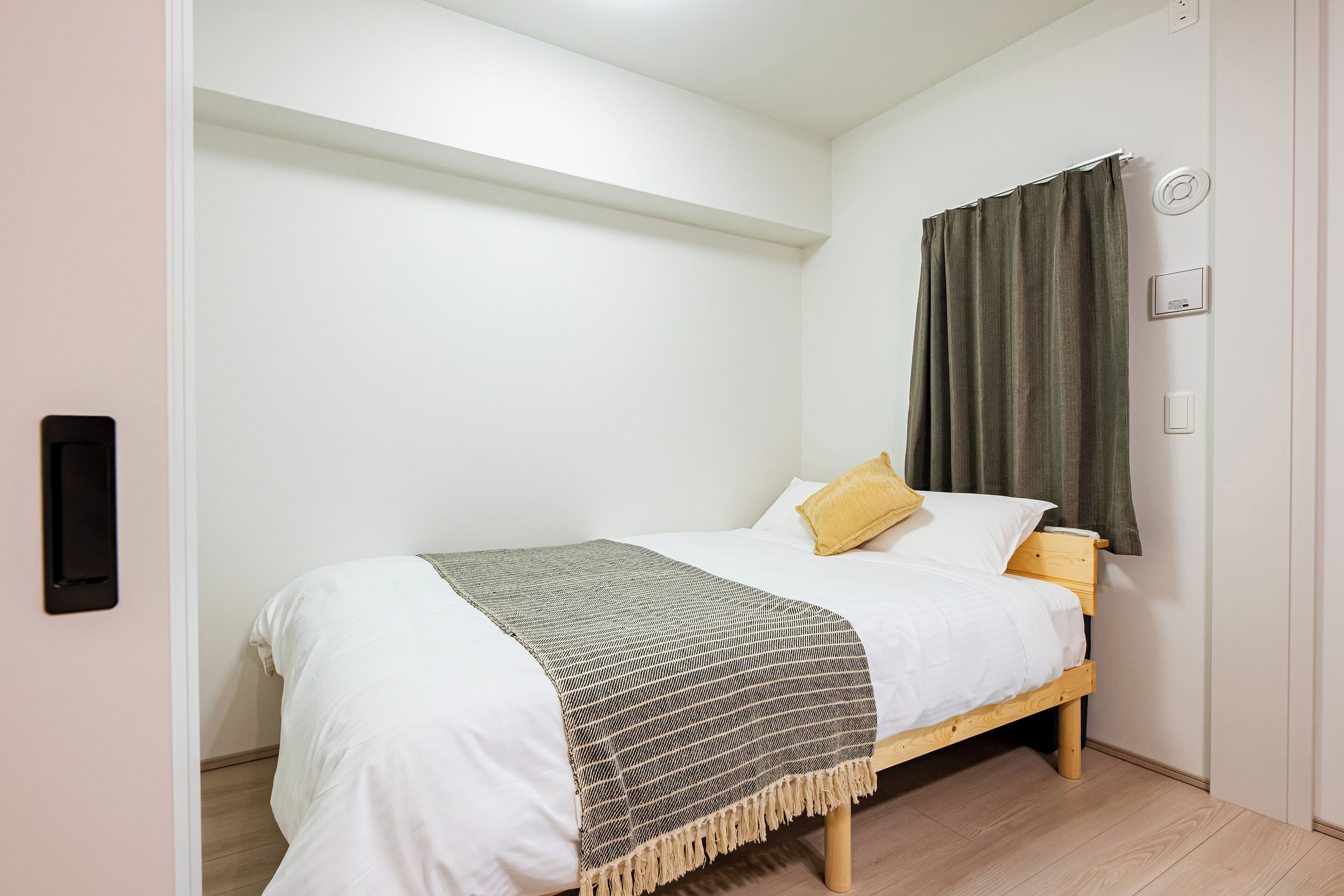 Ein kleines, ordentlich bezogenes Bett mit einer weiß-beigen Bettdecke, einer grau-beigen Streifenwolldecke und zwei Kissen, eines gelb und eines weiß, in einem Zimmer mit weißen Wänden und Holzboden.