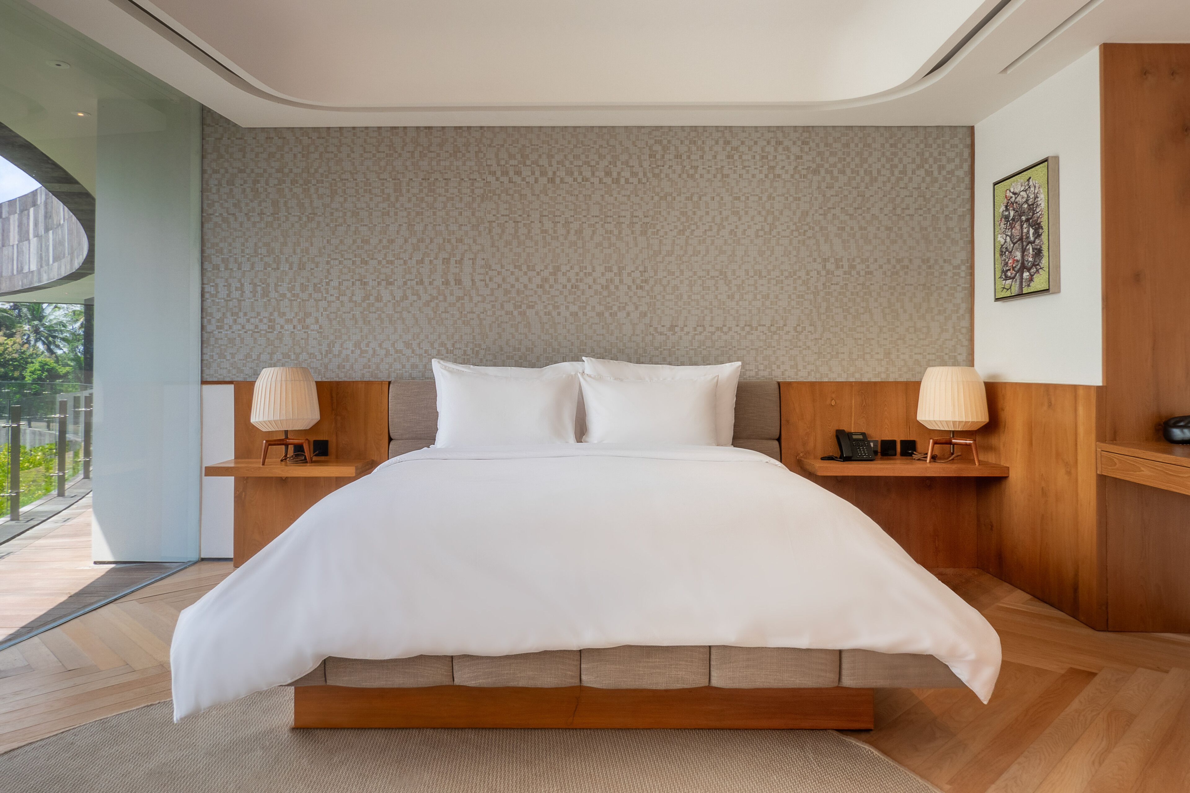 A modern bedroom with a large bed, wooden headboard, bedside lamps, and a view of greenery.