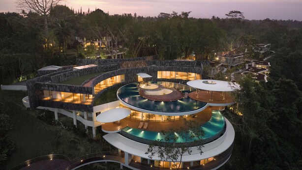 Un complexe hôtelier moderne doté de plusieurs piscines circulaires reliées par des passerelles, entouré d’une végétation luxuriante et adossé à un paysage montagneux.