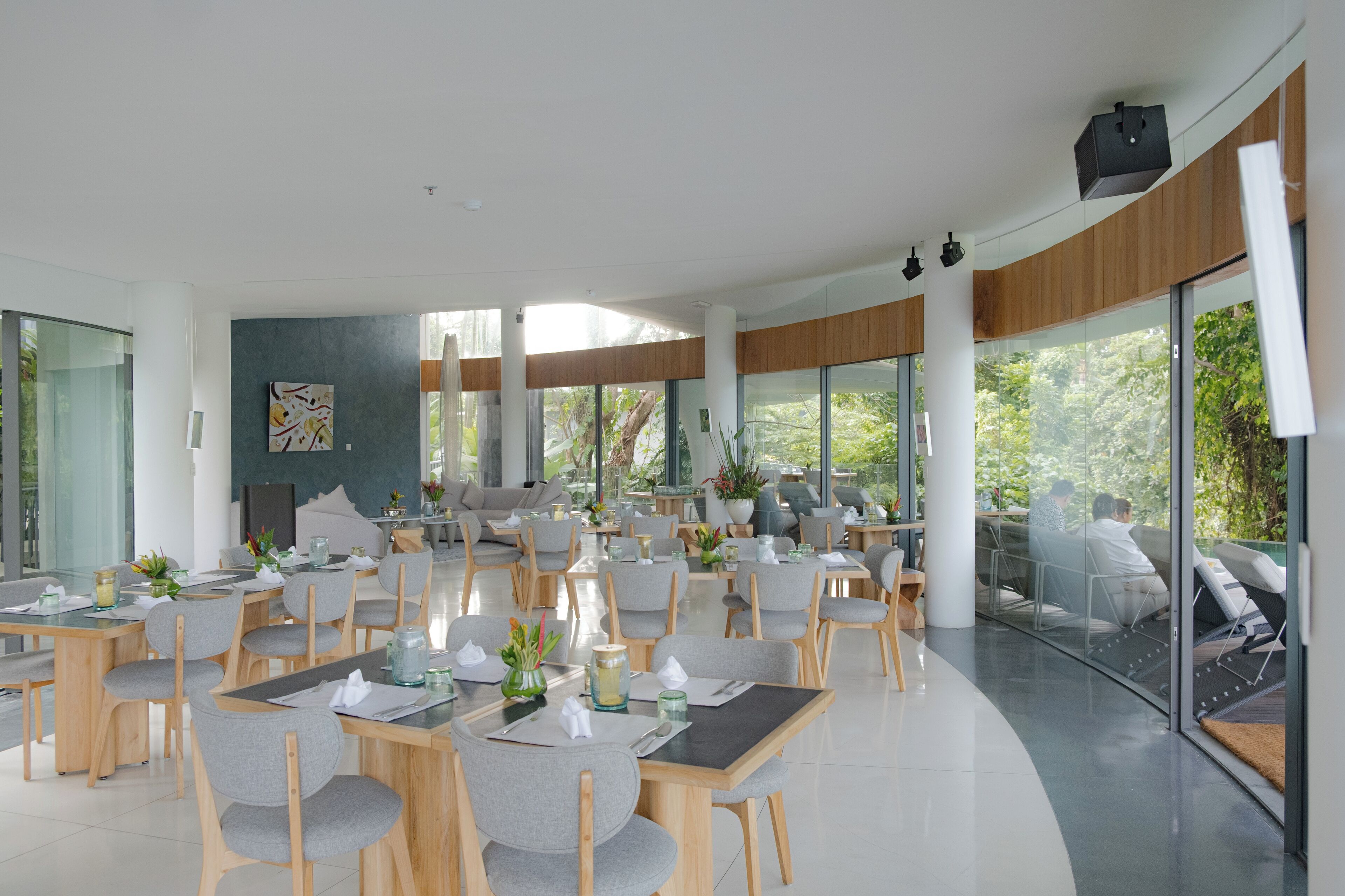 A modern dining area with multiple tables and chairs, large windows offering views of greenery, and a contemporary interior design.