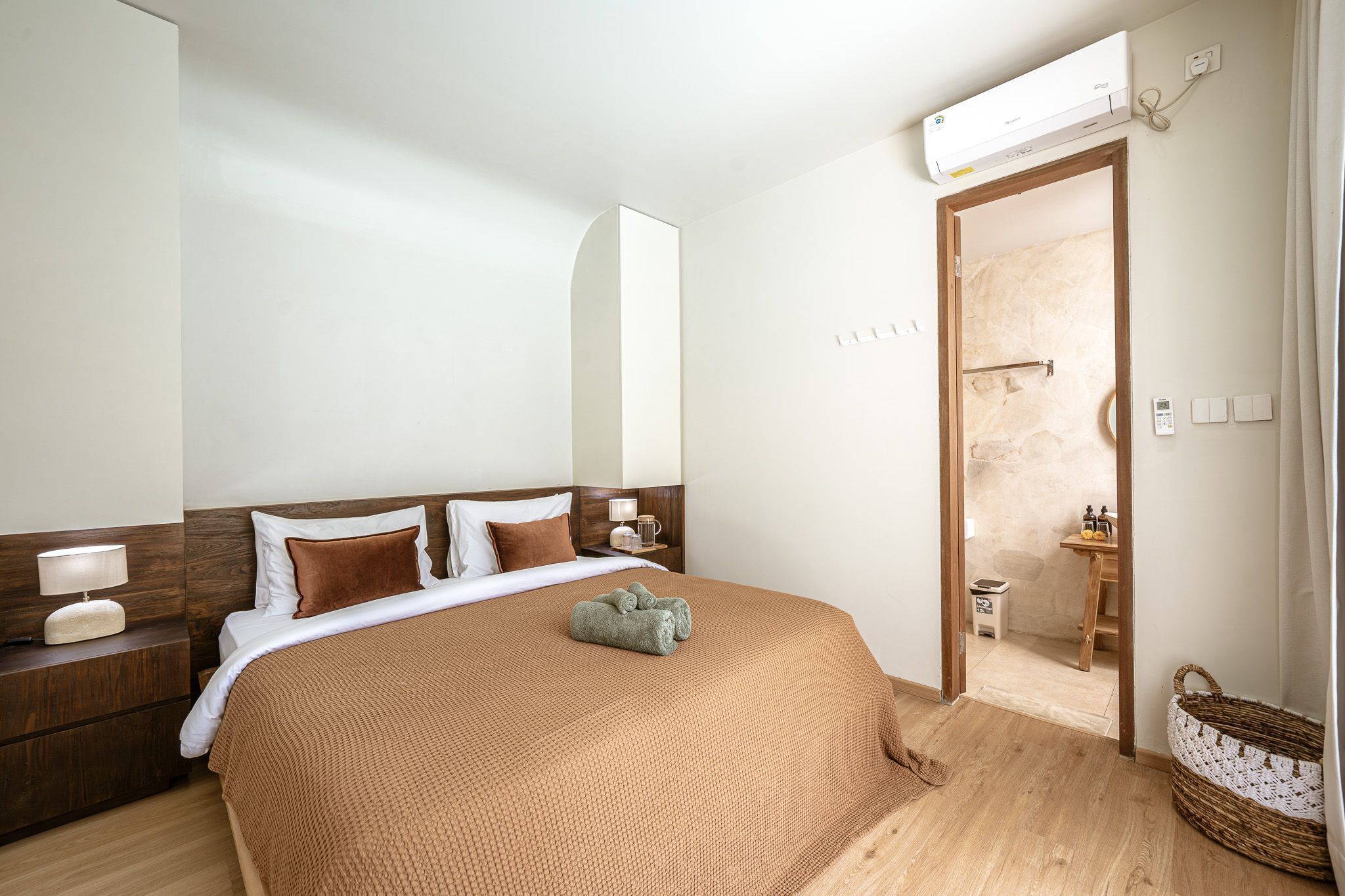 Een hotelkamer met een groot bed, een houten hoofdbord, nachtkastjes met lampen, een klein nachtkastje met een lamp, een wandairconditioner, een houten deur naar een badkamer en een rieten mand op de vloer.