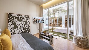 A modern bedroom with a bed, a TV, a painting, and a view of an outdoor area with a tree and a dining table.