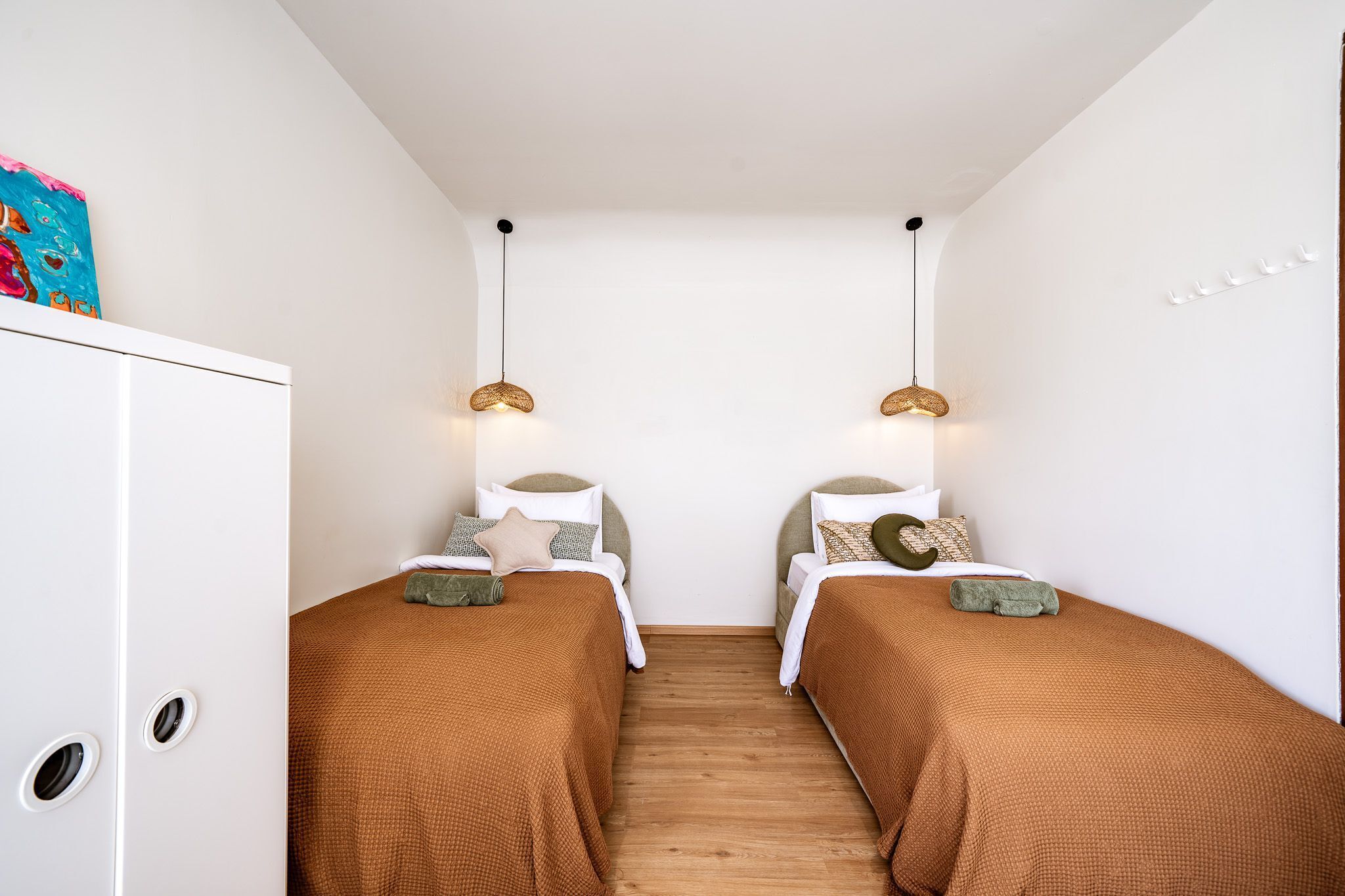 Una habitación pequeña y acogedora con dos camas individuales, cada una con un edredón y almohadas marrones. Hay dos lámparas colgantes en el techo, y un armario blanco a la izquierda.